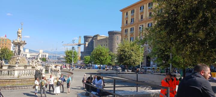 Bustling city square near Castel Nuovo with fountains, historic towers and crowds under blue sky.
