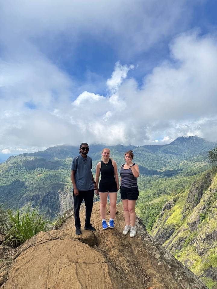 Three hikers stand on a mountaintop viewpoint overlooking lush green valleys beneath dramatic clouds.