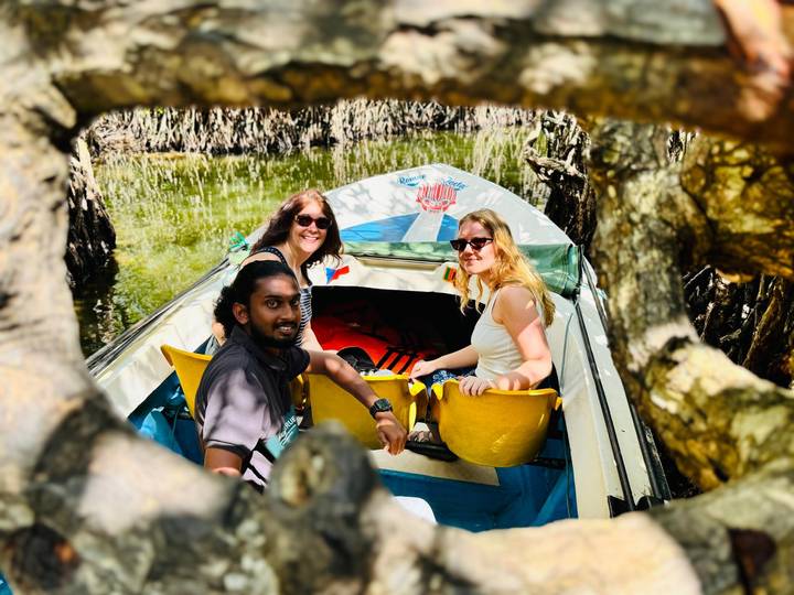 Three travelers smile from a small boat paddling through lush mangrove tunnels framed by twisted branches.