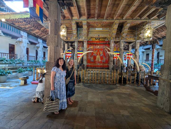 Two visitors stand inside the richly decorated Temple of the Tooth surrounded by ivory tusks and murals.
