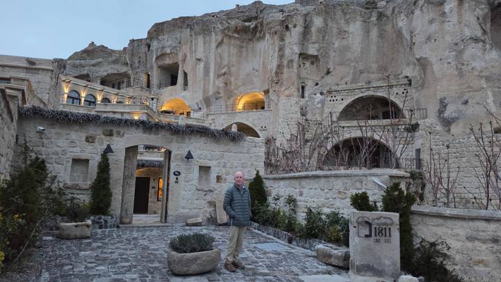 Traveler stands in the courtyard of a cave-style stone hotel carved into dramatic cliffs, warm lights glowing at dusk.