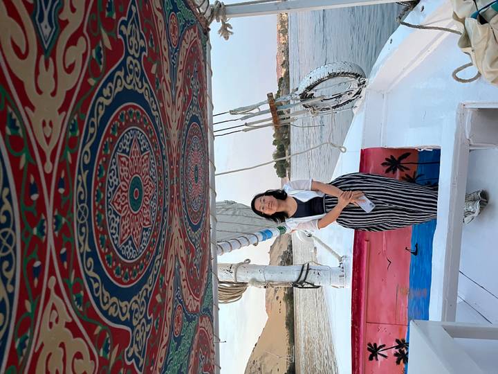 A woman stands on the bow of a felucca under a patterned canopy sailing the Nile at dusk.