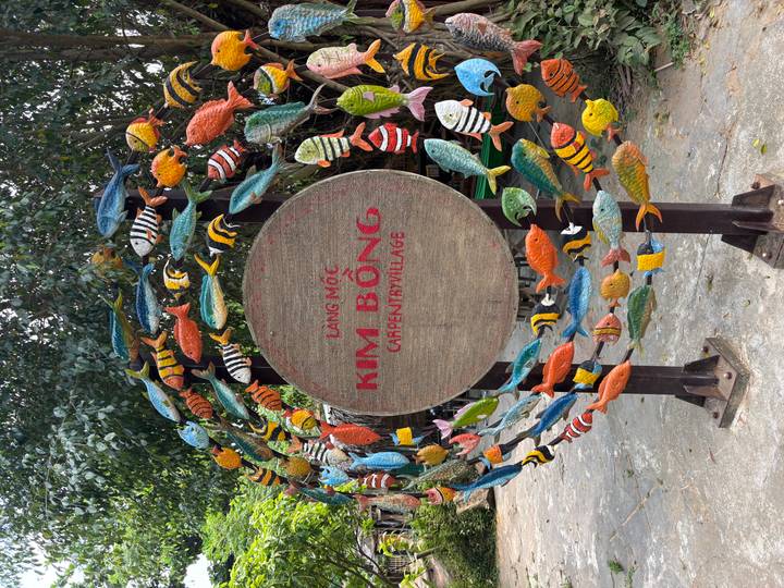 Colorful fish carvings form a round sign reading 'Kim Bồng Carpentry Village' in a leafy courtyard.