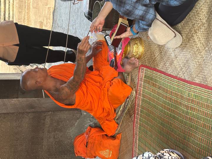 Orange-robed Buddhist monk ties a blessing thread on a visitor inside an Angkor temple alcove.