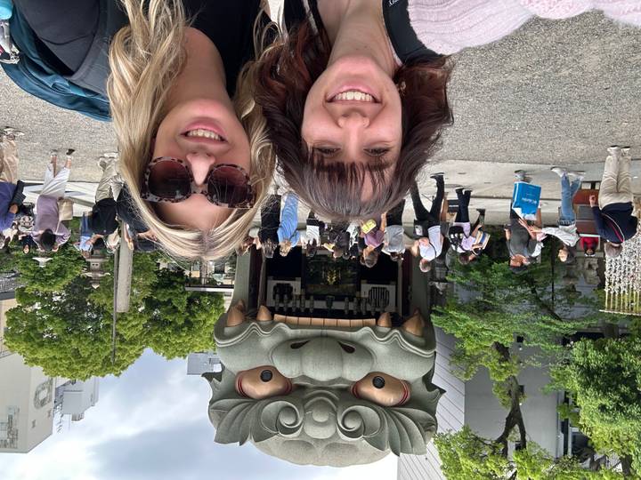 Two travelers smile before giant open-mouthed lion head shrine as crowds wander nearby.