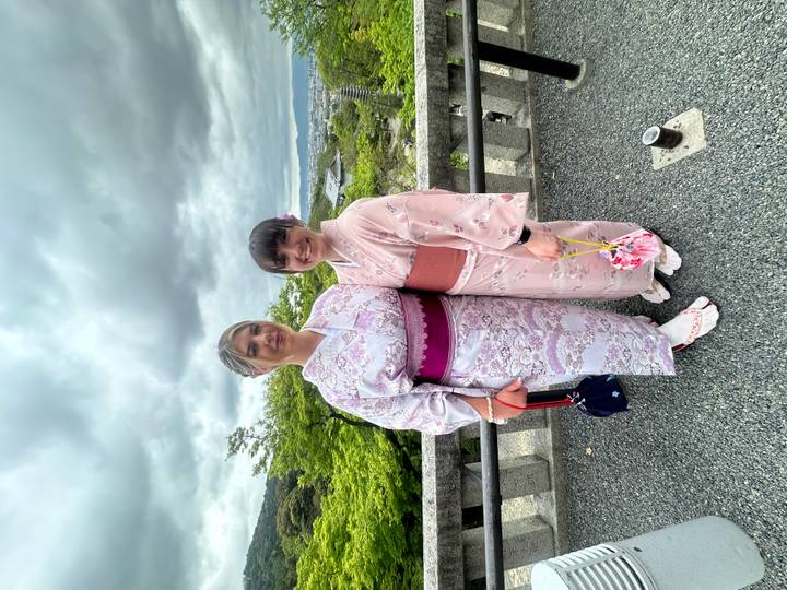 Two women wearing pastel kimonos pose on viewpoint overlooking cityscape.
