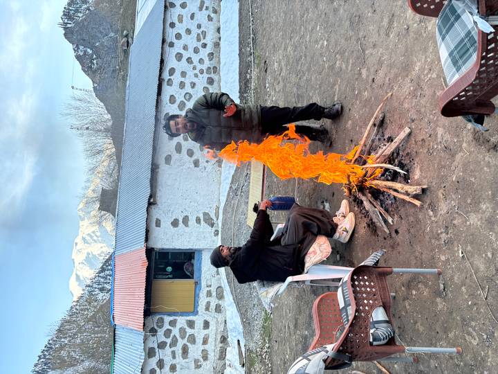 Two men warming by a tall orange campfire flame outside a rustic lodge.