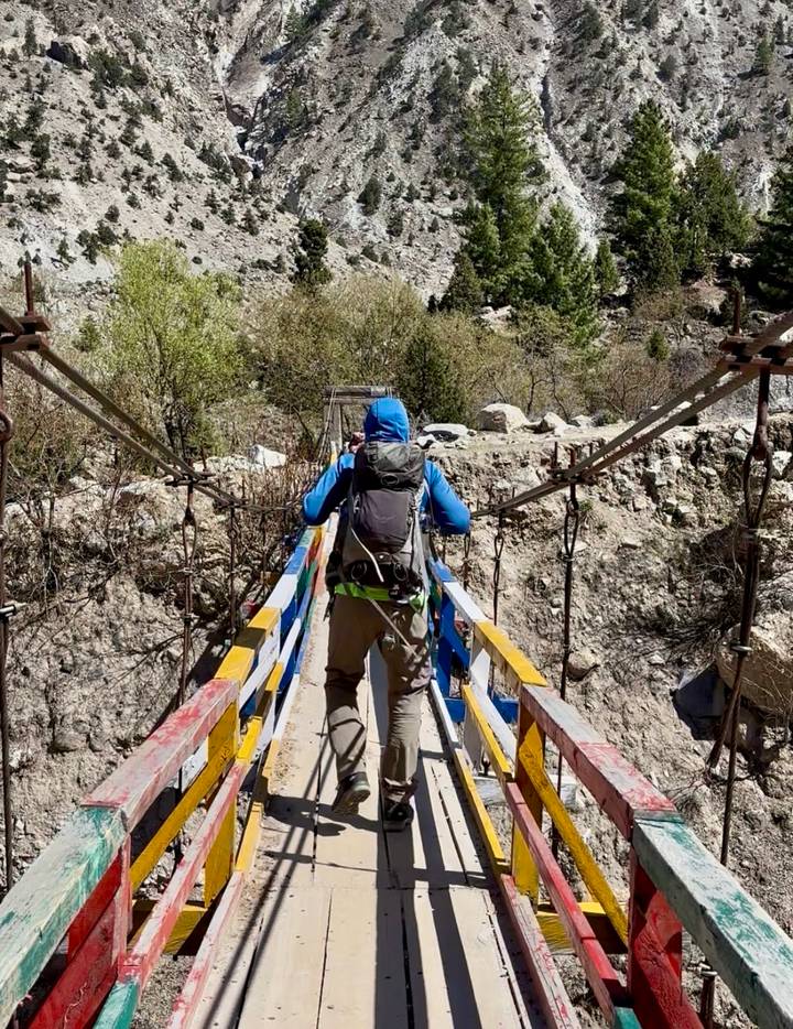 Back view of a trekker crossing a colorful wooden suspension bridge over a rocky gorge.