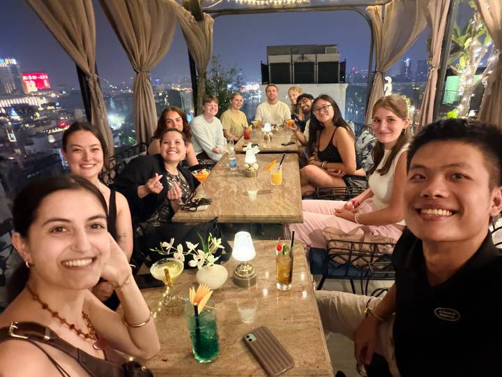 Large group of friends enjoying cocktails at a rooftop bar overlooking Hanoi’s night skyline.