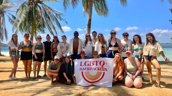 LGBTQ+ backpackers group poses on a tropical beach under palm trees with a rainbow banner.