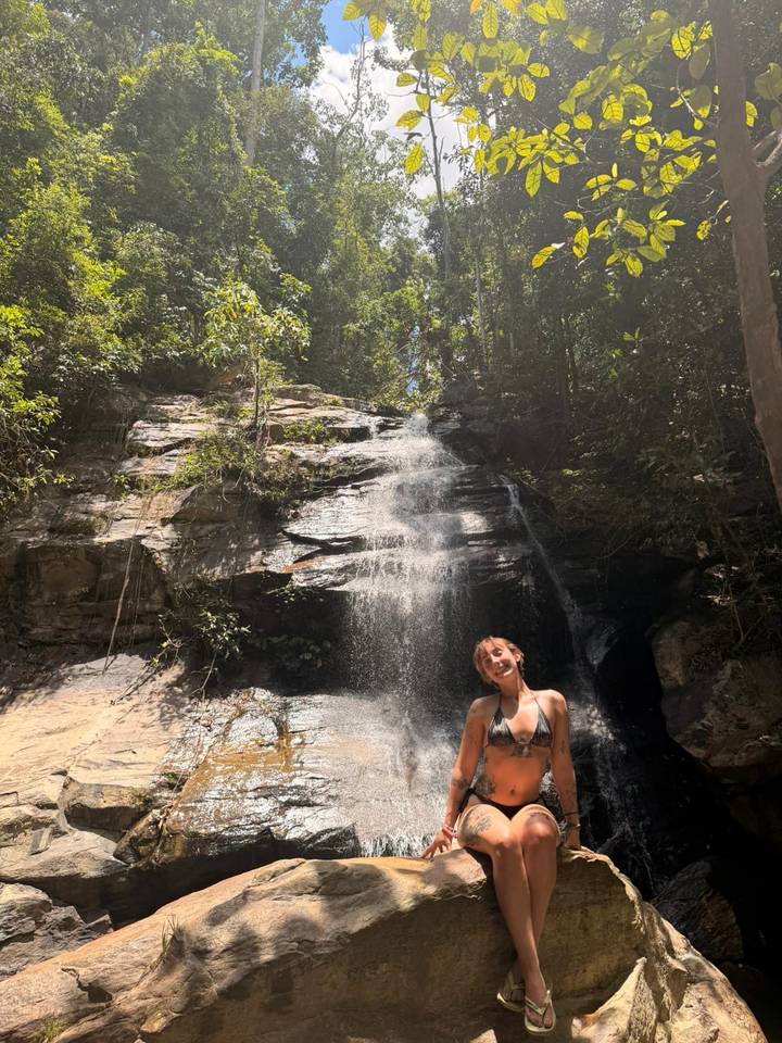 Woman stands smiling at the base of a jungle waterfall with sunlight streaming through the trees.