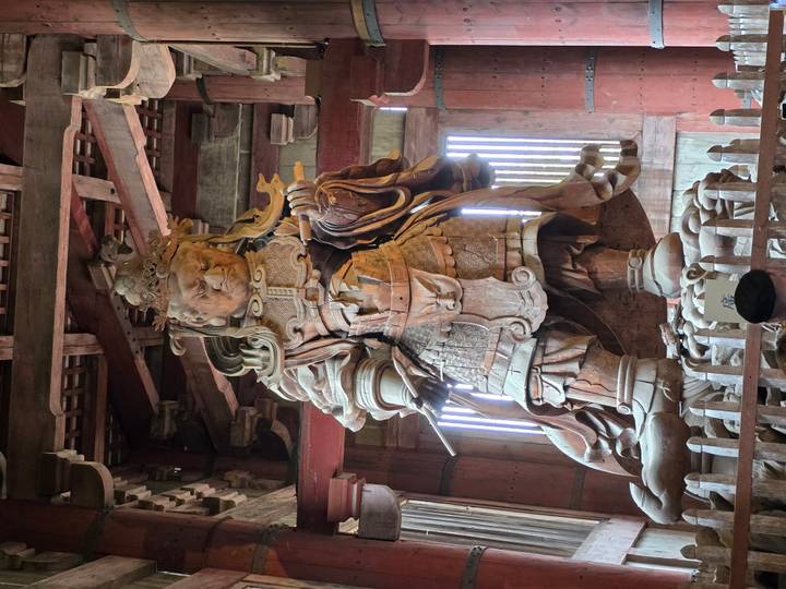 Massive wooden warrior statue stands inside a dim temple hall with beams overhead.