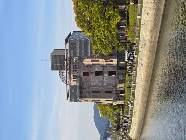 The A-Bomb Dome ruins stand beside a calm river lined with green trees in Hiroshima.