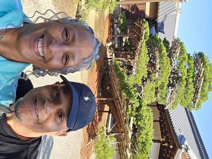 Close selfie of a smiling couple with a sculpted pine tree and traditional roof in the background.