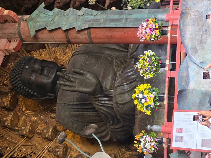 Giant bronze Buddha statue raises a hand in blessing inside Todaiji Temple, flanked by floral offerings.