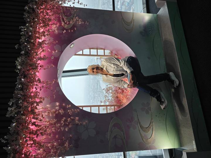 Woman poses inside a circular window display decorated with pink blossoms overlooking Tokyo skyline.