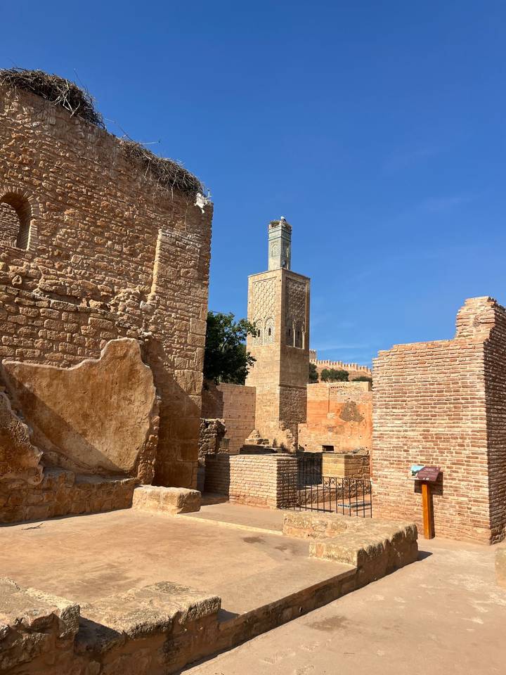 Sunlit ancient brick walls and a slender minaret rise amid archaeological ruins against a clear sky.