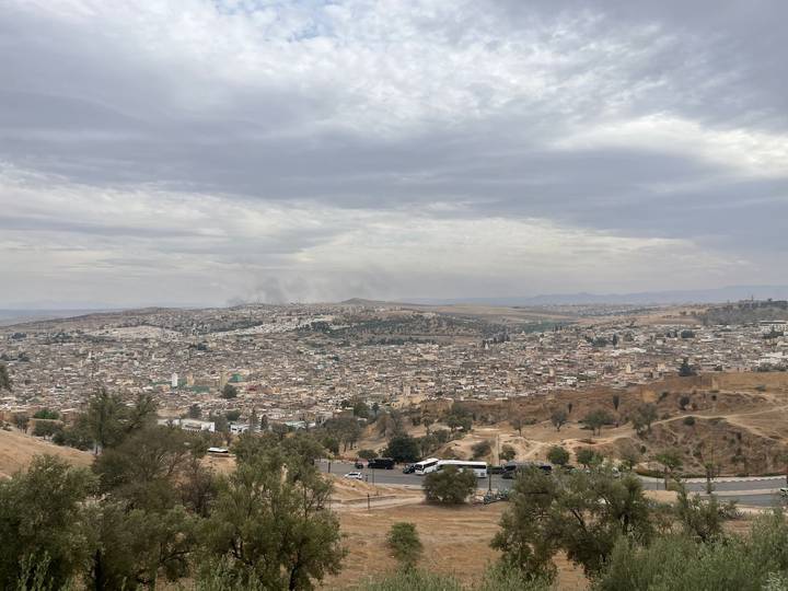 Wide, hazy panorama of Fes spreads beneath an overcast sky, with smoke plumes rising in the distance.
