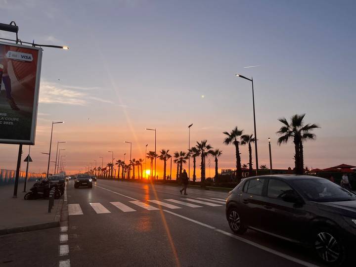 Cars and palm trees line a seaside boulevard as the sun sets in a fiery orange glow.