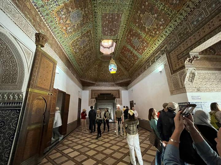 Tourists explore an ornately decorated hall with detailed mosaic ceilings in Morocco.