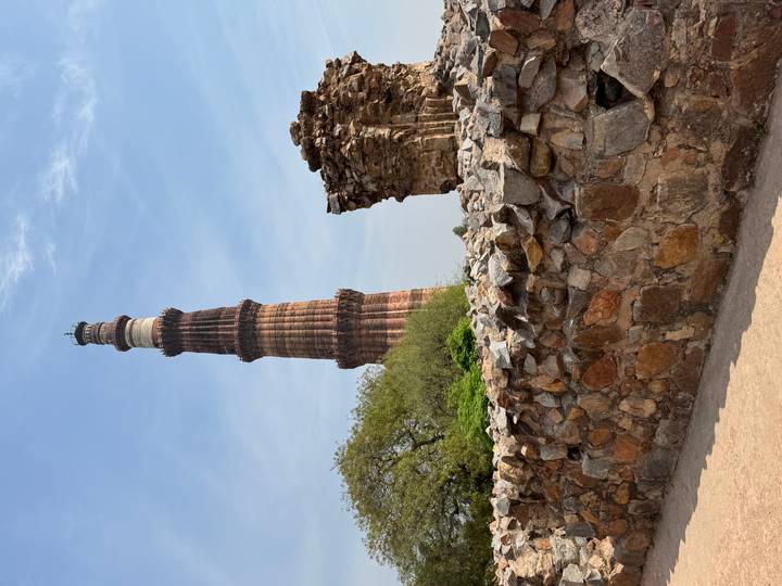 Tall red-sandstone Qutub Minar rises above ancient stone ruins and trees against a blue sky.
