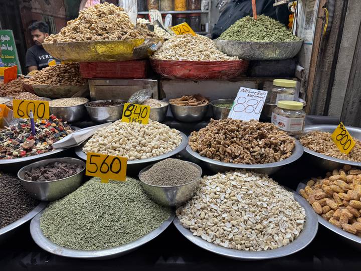 Assorted nuts, seeds and spices displayed in metal bowls with handwritten price tags at a market.