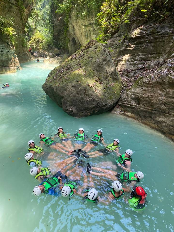 Adventurers wearing helmets and life vests float in a circle in turquoise canyon waters beneath a large boulder.