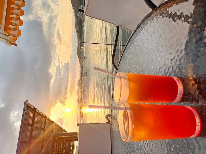 Two vibrant cocktails on a glass table overlook boats and a colorful sunset on the bay.