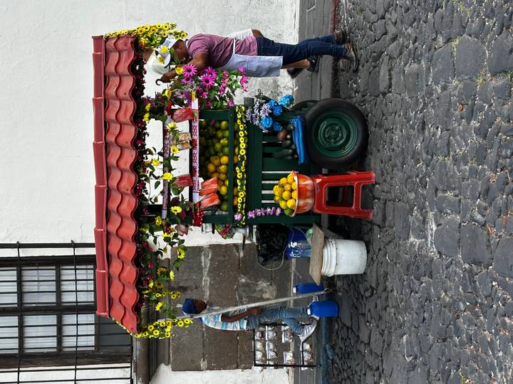 Street vendor prepares fresh fruit juices at a colorful cart decorated with flowers in Antigua's cobblestone street.