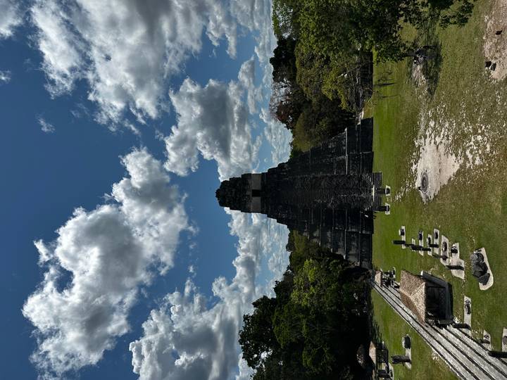 Stepped Mayan pyramid rises amid jungle clearing in Tikal National Park under dramatic cloud-filled skies.