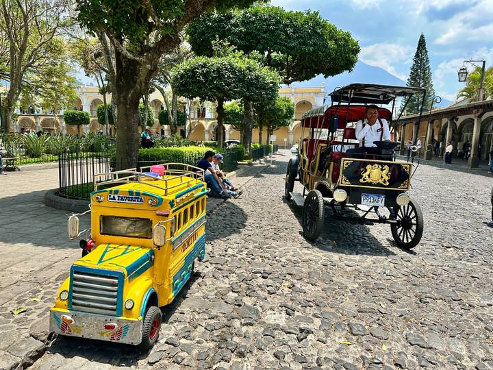 Colorful chicken bus toy and horse carriage share a sunlit cobblestone plaza in historic Antigua.