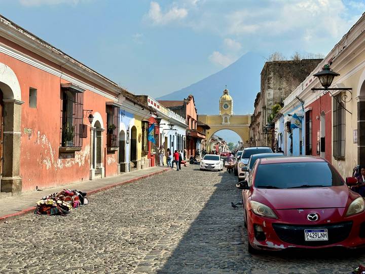 Busy colonial street in Antigua with colorful facades, parked cars, and distant archway beneath a hazy volcano.