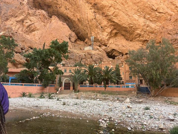 Clay-colored buildings and tall palms nestle against sheer orange cliffs beside a dry riverbed at Todra Gorge.