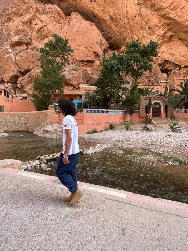 A visitor pauses near a shallow stream with the ochre walls of Todra Gorge rising behind.