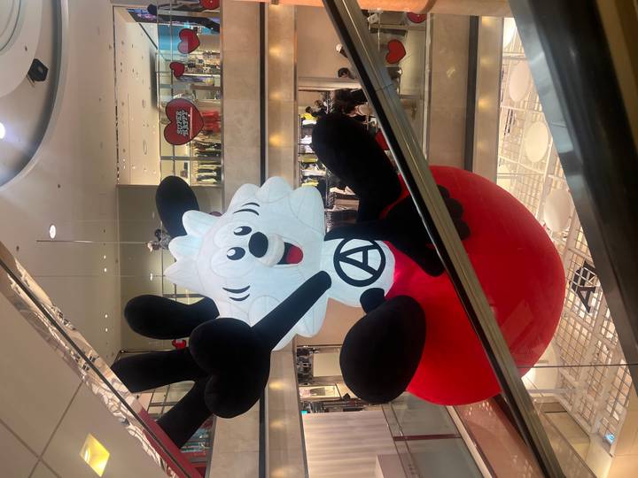 Large cartoon-style inflatable character hanging inside a multi-level shopping mall filled with shoppers.