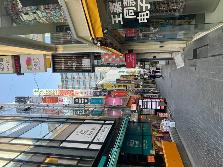 Narrow shopping street lined with tall signboards and shopfronts in a busy Seoul district.