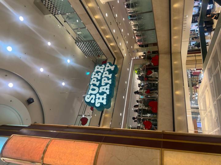 Interior of a shopping mall with a large ‘SUPER HAPPY’ sign hanging above crowds on multiple levels.