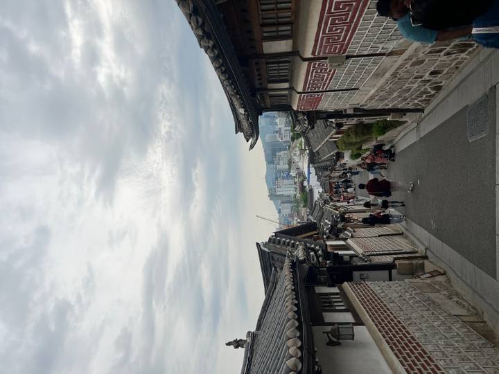 Traditional hanok alley descending towards modern Seoul skyline under a cloudy sky with tourists walking.