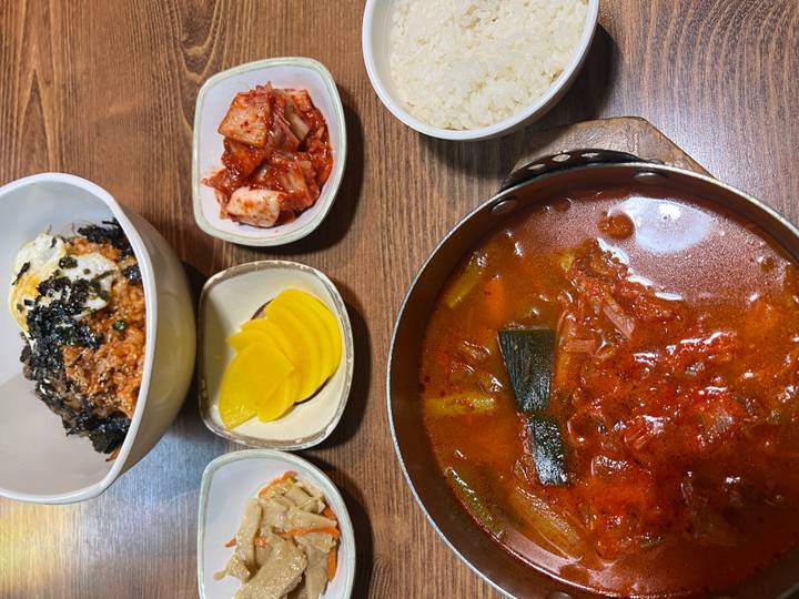 Korean meal featuring a bubbling red kimchi stew, rice, pickles and side dishes on a wooden table.