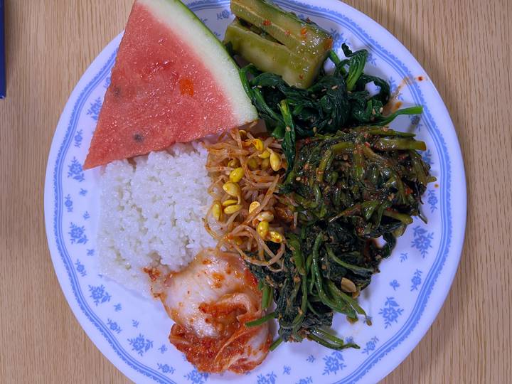 Close-up plate with rice, kimchi, greens, bean sprouts and a slice of watermelon on a wooden table.