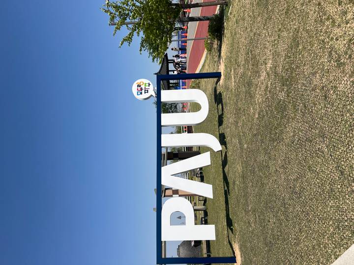 Outdoor sign spelling PAJU in a grassy park area under a clear sky with visitors nearby.