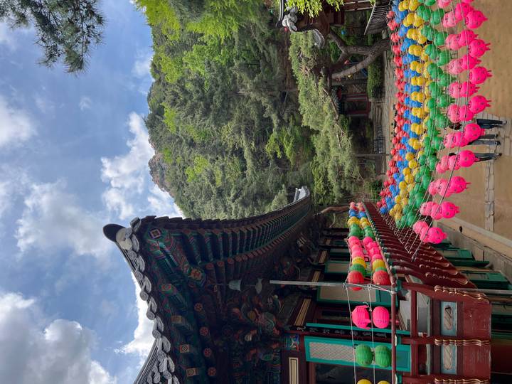 Vibrant lanterns strung in front of a mountain temple with forested cliffs rising behind under a partly cloudy sky.
