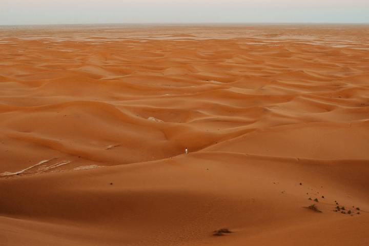 Vast orange dunes with a lone figure standing small against the sweeping desert landscape.