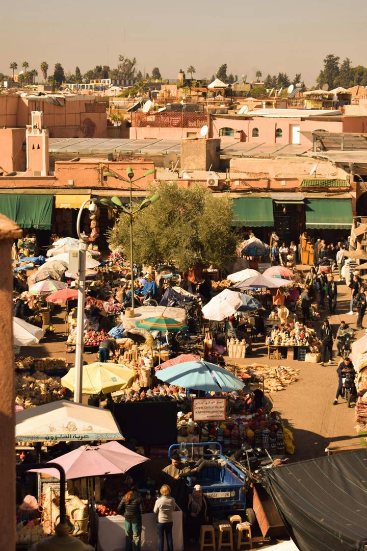 Bustling outdoor market packed with colorful umbrellas, stalls of goods, and crowds of shoppers in a sunny plaza.