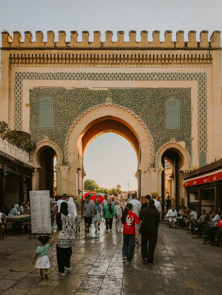 Crowds walk beneath the ornate green-tiled Bab Bou Jeloud gate leading into the medina at sunset glow.