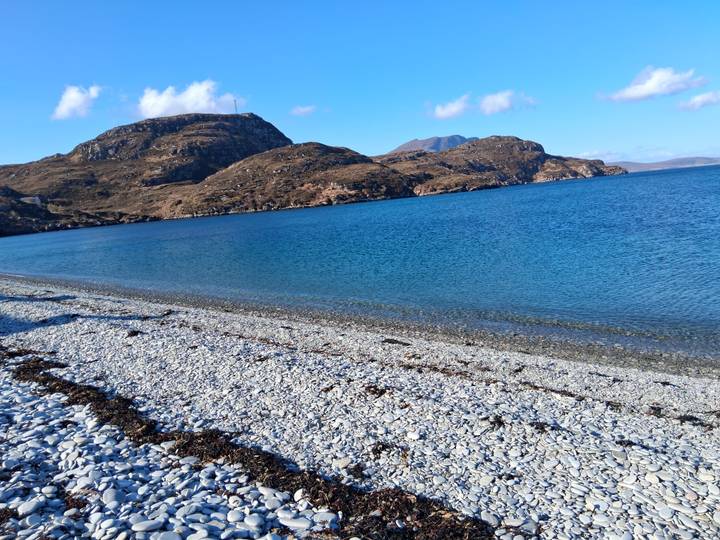 Pebble beach lapped by crystal clear turquoise water with rugged hills in the background on a sunny day.