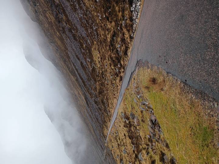 Narrow single-track road winds up a mist-shrouded Highland mountainside on a rainy day.