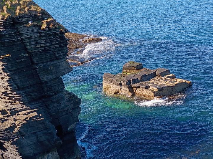 Rocky sea stack rises from vibrant turquoise waters along Scotland’s dramatic cliff coast.