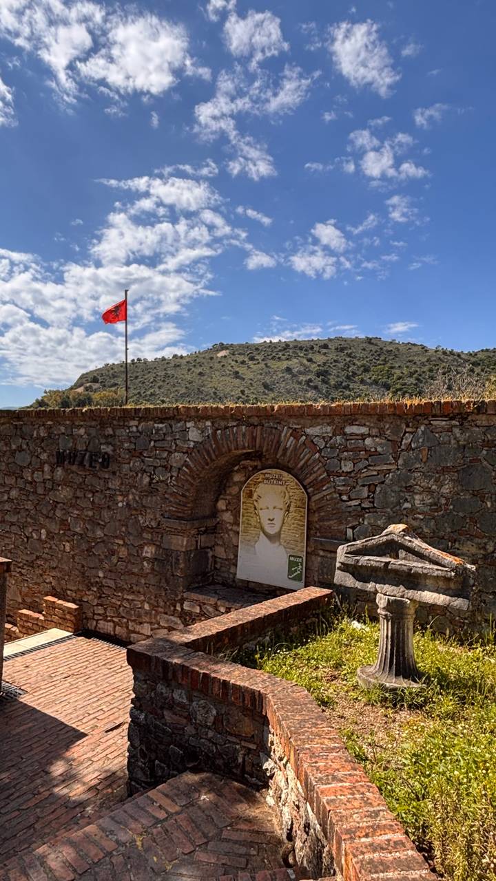 Stone fort wall with Albanian flag and hill backdrop near historic entrance arch.