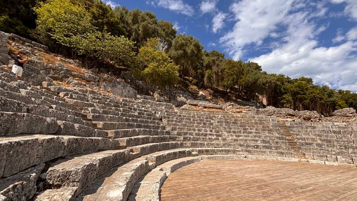 Stone amphitheater seating curves through forested hillside under blue sky.